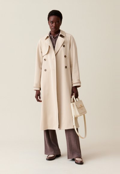 Cappotto beige in stile trench con bottoni doppi, ampi risvolti e una vestibilità rilassata, abbinato a pantaloni marroni a coste e una piccola borsa bianca.