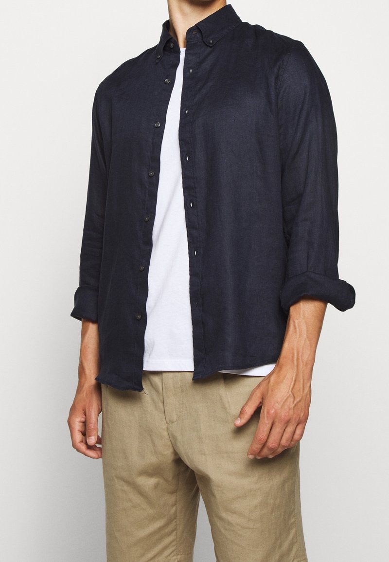 Homme portant une chemise boutonnée bleu foncé sur un t-shirt blanc, associé à un short beige, se tenant devant un fond uni.