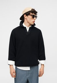Jeune homme portant un pull en tricot noir, une chemise blanche, un jean bleu, une casquette beige portée à l'envers et des lunettes de soleil foncées, regardant sur le côté.