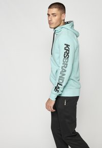 Sudadera verde menta con diseño de texto negro en la manga, capucha ajustable con cordones y bolsillo frontal, combinada con pantalones negros con detalle del logo.
