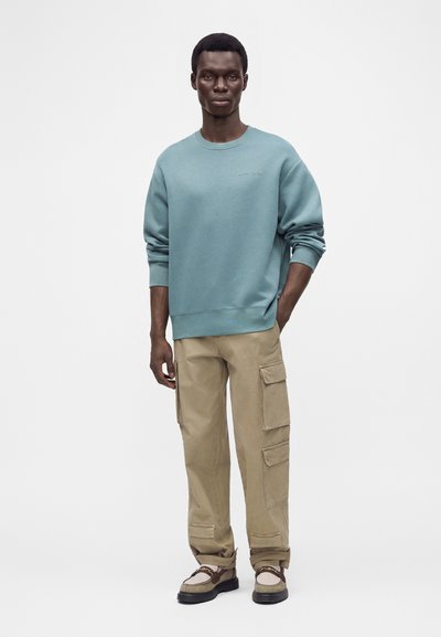 Homme debout avec une main dans la poche, portant un sweat-shirt bleu, un pantalon cargo beige et des chaussures marron et blanches sur un fond uni.