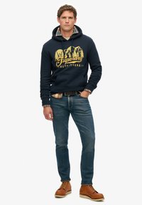 Marineblå hættetrøje med guld bjerggrafik og "Superdry Outfitters" tekst, lagdelt over en ternet skjorte, sammen med denim jeans og brune støvler.
