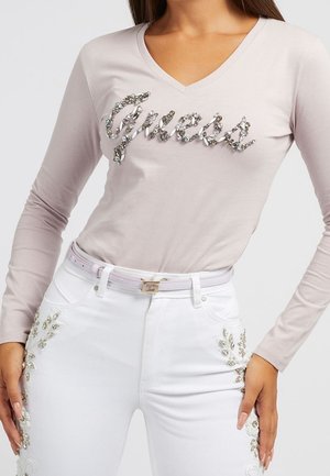 Kvinde iført sart lyserød langærmet top med juvelbesat "Guess" logo og hvide jeans med blomsterperleudsmykninger på siderne.
