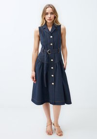 Jeans-Minikleid ohne Ärmel mit tailliertem Gürtel, Knopfleiste und ausgestelltem Rock. Dunkelblaue Farbe mit kontrastierender Naht und goldenen Beschlägen.