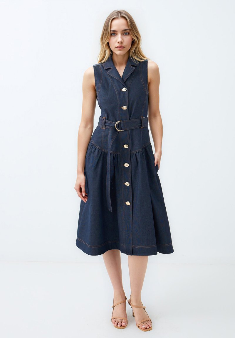 Robe sans manches en denim avec une taille ceinturée, un devant boutonné et une jupe évasée. Couleur bleu foncé avec des coutures contrastantes et des accessoires en or.