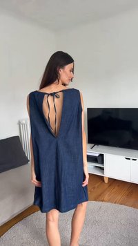 Γυναίκα μέσα σε εσωτερικό χώρο που φοράει αμάνικο σκούρο μπλε φόρεμα με βαθύ V στην πλάτη δεμένο στην κορυφή, στεκόμενη κοντά σε γκρι καναπέ και λευκό έπιπλο τηλεόρασης με μαύρη οθόνη.