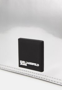 Borsa a tracolla metallica argento con patch logo nero; presenta una texture liscia, forma rettangolare e dettagli di cuciture bianche.