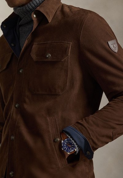 Καφέ σατέν μπουφάν με μπροστινές τσέπες, κουμπιά με επικόλληση και υφασμένο γιακά. Φορεμένο πάνω από ένα γκρι πλεκτό ζιβάγκο με μπλε λεπτομέρεια μανσετών.