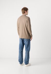 Camisa bege de manga longa feita de material suave, combinado com jeans azuis de corte largo. Costura notável e um design simples. Ténis brancos.