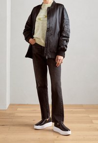 Veste bomber noire avec poignets côtelés, pull texturé vert clair avec logo, jean foncé, et chaussures slip-on foncées avec semelles blanches.