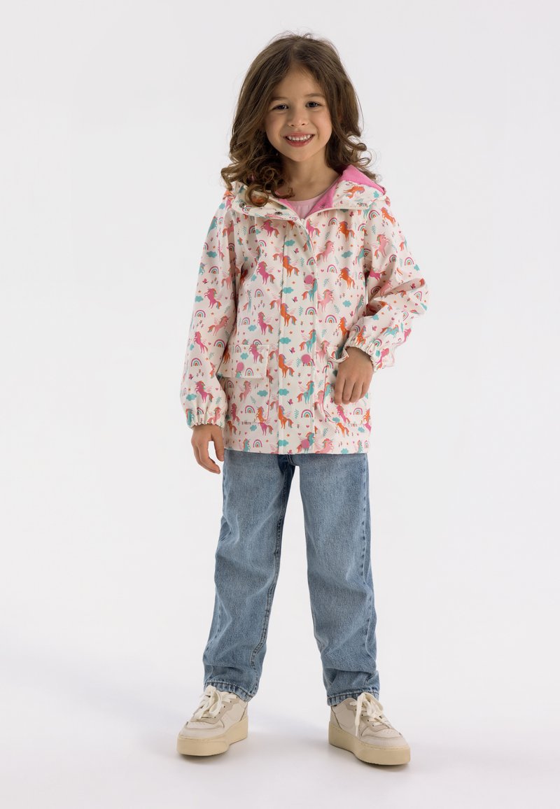 Giacca impermiabile rosa chiaro con motivi di unicorni e arcobaleni, dotata di cappuccio, polsini elasticizzati e tasche, abbinata a jeans blu e sneaker.