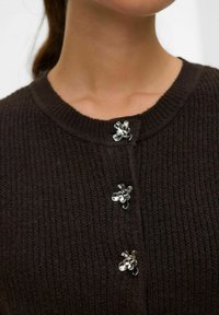 Hnědý pletený cardigan s kulatým výstřihem, který vpředu zdobí tři kovové knoflíky ve tvaru květin pro zapínání a detail.