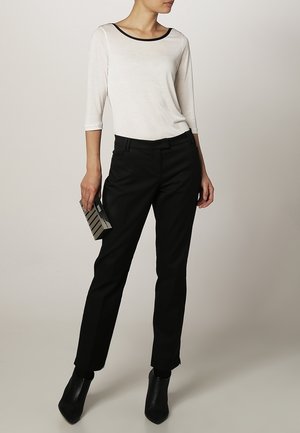 Vrouw draagt een wit topje met zwarte bies, zwarte broek en zwarte enkellaarzen, en houdt een zwarte en zilveren clutch vast.