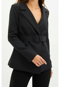 Musta bleiseri, jossa on vartalonmyötäinen malli, terävät kaulukset ja vastaava vyö, jossa on pyöreä solki. Sileä pinta ja räätälöity istuvuus.