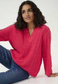 Pull en tricot rose vif avec un col en V, présentant un motif texturé et des détails en maille ouverte, associé à un jean en denim bleu.