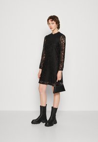 Black lace dress with long, semi-sheer sleeves, scalloped neckline, and floral pattern. Paired with black chunky ankle boots and a small handbag.