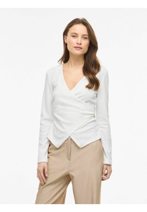 Kobieta o długich brązowych włosach, ubrana w białą bluzkę z długim rękawem w stylu kopertowym oraz beżowe spodnie z wysokim stanem, stojąca na jednolitym tle.