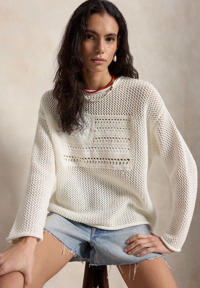Νεαρή γυναίκα με μακριά σκούρα μαλλιά που φοράει ένα λευκό πουλόβερ ανοιχτής πλέξης με σχέδιο και ανοιχτόχρωμα τζιν σορτς κάθεται σε ένα σκαμπό.