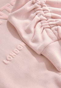 Felpa rosa chiaro con tessuto testurizzato, dettagli con maniche arricciate e logo "LONDON" in rilievo. Materiale morbido e stretchable con polsini a coste.
