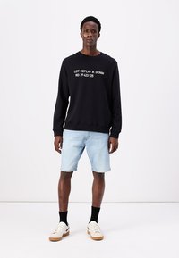 Sweatshirt preto com texto branco, calções de ganga azul claro e ténis bege com acabamentos brancos. Design simples, corte descontraído e gola redonda.