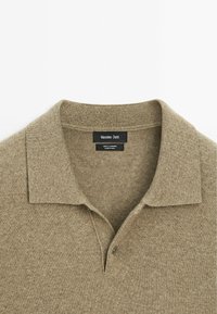 Beige Kaschmir-Polohemd mit klassischem Kragen, das über eine subtile Textur und ein kleines Knopfdetail am Hals verfügt. Markenlabel innen sichtbar.