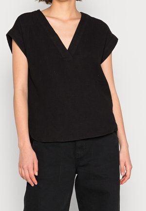 Vrouw die een losse zwarte V-hals T-shirt met korte mouwen en een zwarte broek draagt, staand met ontspannen armen tegen een effen achtergrond.