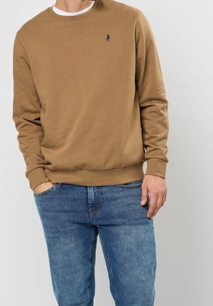 Man draagt een bruine longsleeve trui met klein geborduurd logo en blauwe spijkerbroek, handen in zakken, tegen een effen achtergrond.