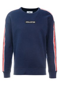 Sweat à capuche bleu marine en coton, avec des rayures latérales rouges et blanches, col rond, et un logo rectangulaire noir "HOLLISTER" sur le devant.