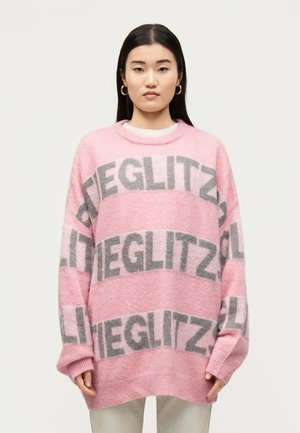 Femme portant un pull ample rose avec un motif de texte gris « GLITZ », associé à un pantalon de couleur claire, debout devant un fond blanc uni.