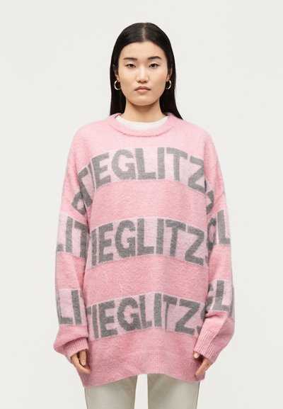 Femme portant un pull ample rose avec un motif de texte gris « GLITZ », associé à un pantalon de couleur claire, debout devant un fond blanc uni.