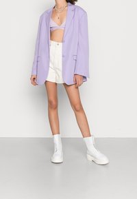 Blazer oversized lavande, crop top blanc, shorts blancs taille haute et bottines blanches à lacets avec semelles épaisses.