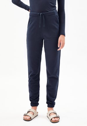 Person trägt marineblaue Jogginghosen mit elastischen Bündchen und beige Schnallensandalen, steht vor weißem Hintergrund.