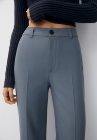 Op maat gemaakte grijze broeken met een vlakke voorkant, zijzakken en een knoopsluiting. Gemaakt van een gladde stof met een subtiele glans.