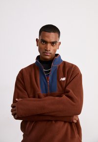 Felpa a mezza zip in pile marrone con colletto blu navy, logo bianco ricamato e maniche lunghe. Texture morbida, vestibilità rilassata e blocchi di colore a contrasto.