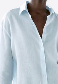 Camicia in lino azzurro chiaro con abbottonatura frontale, colletto classico e vestibilità rilassata. Presenta maniche lunghe e un tessuto testurizzato e traspirante.