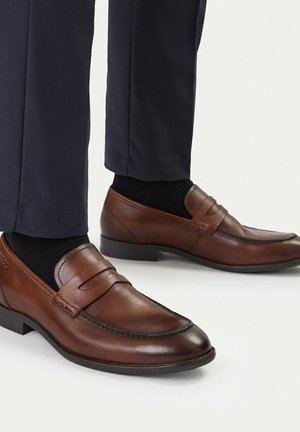 Braune Lederloafers, getragen mit schwarzen Socken und marineblauen Hosen, seitlich gezeigt vor einem schlichten hellen Hintergrund.
