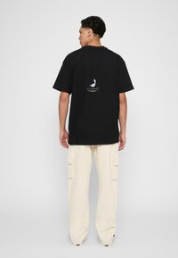 Sort bomulds-T-shirt med korte ærmer, der har et grafisk detalje på bagsiden. Kombination med beige cargo bukser med sidelommer.