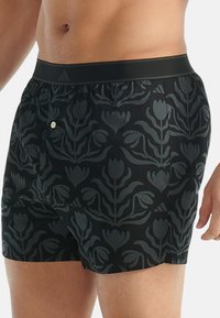 Pantaloni da bagno neri con un motivo floreale a tono, vita elastica e piccolo dettaglio metallico; realizzati in tessuto leggero e liscio.