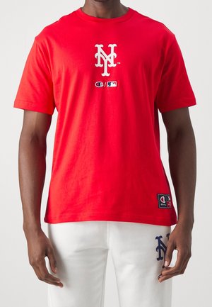 Mężczyzna w czerwonej koszulce z białym logo "NY" oraz logo Champion i MLB, w połączeniu z białymi spodniami z granatowym logo "NY".
