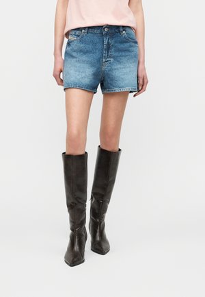 Persoană îmbrăcată cu șorturi albastre din denim, un top roz deschis și cizme maro închis din piele până la genunchi, stând în fața unui fundal simplu.