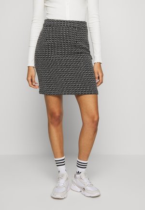 Kobieta ubrana w czarno-białą mini-spódniczkę w kratę, biały top z długim rękawem, białe skarpety Adidas z czarnymi paskami oraz białe trampki.