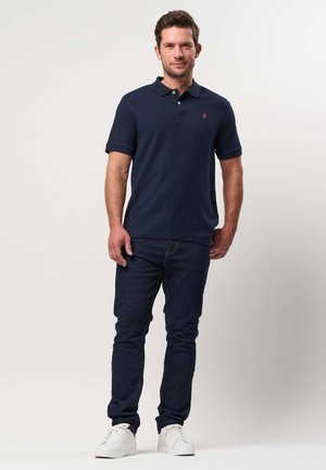 Vīrietis stāv, valkājot tumši zilu polo kreklu, tumši zilas džinsas un baltus sporta apavus pret vienkāršu gaišu fonu.