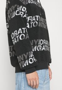 Sweat à capuche en polaire noire avec un motif de texte blanc et gris sur toute la surface, porté avec un jean bleu clair, vu de côté.