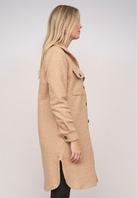 Beige kabát z vlněné směsi s texturovanou látkou, zapínání na knoflíky, dvě náprsní kapsy a boční rozparek, nošený s černými legínami.