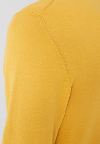 Gros plan sur l'épaule et la partie supérieure de la manche d'un pull en maille jaune moutarde avec des détails de couture visibles.