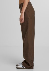 Braune, weite Hosen aus robustem Stoff. Verfügt über zwei Gesäßtaschen und eine hohe Taille. Kombiniert mit weißen und grauen Sneakers.