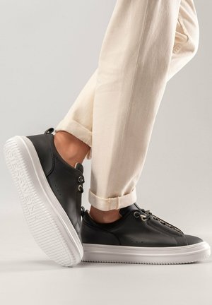 Schwarze Ledersneaker mit glatter Textur, weißer Gummisohle und runden Schnürsenkeln. Kombiniert mit hellen, hochgekrempelten Hosen, die den Knöchel zeigen.