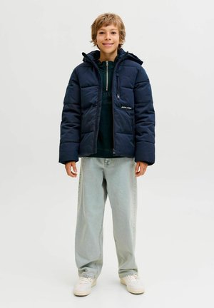 Jauns cilvēks stāv un smaida, uzvilcis tumši zilu pūkaino jaku, gaiši zilus džinsus un baltus sneakers uz viendabīga fona.