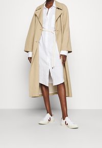 Beige lange jas over een witte blousejurk met knopen en een tailleband, gecombineerd met witte sneakers met rode accenten. Gladde texturen overal.