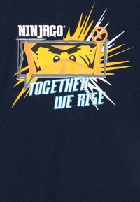 Morska majica s Ninjago temom koja ima žutu grafiku lica s maskom, okružena dizajnom eksplozije. Tekst glasi "Zajedno se uspinjemo."